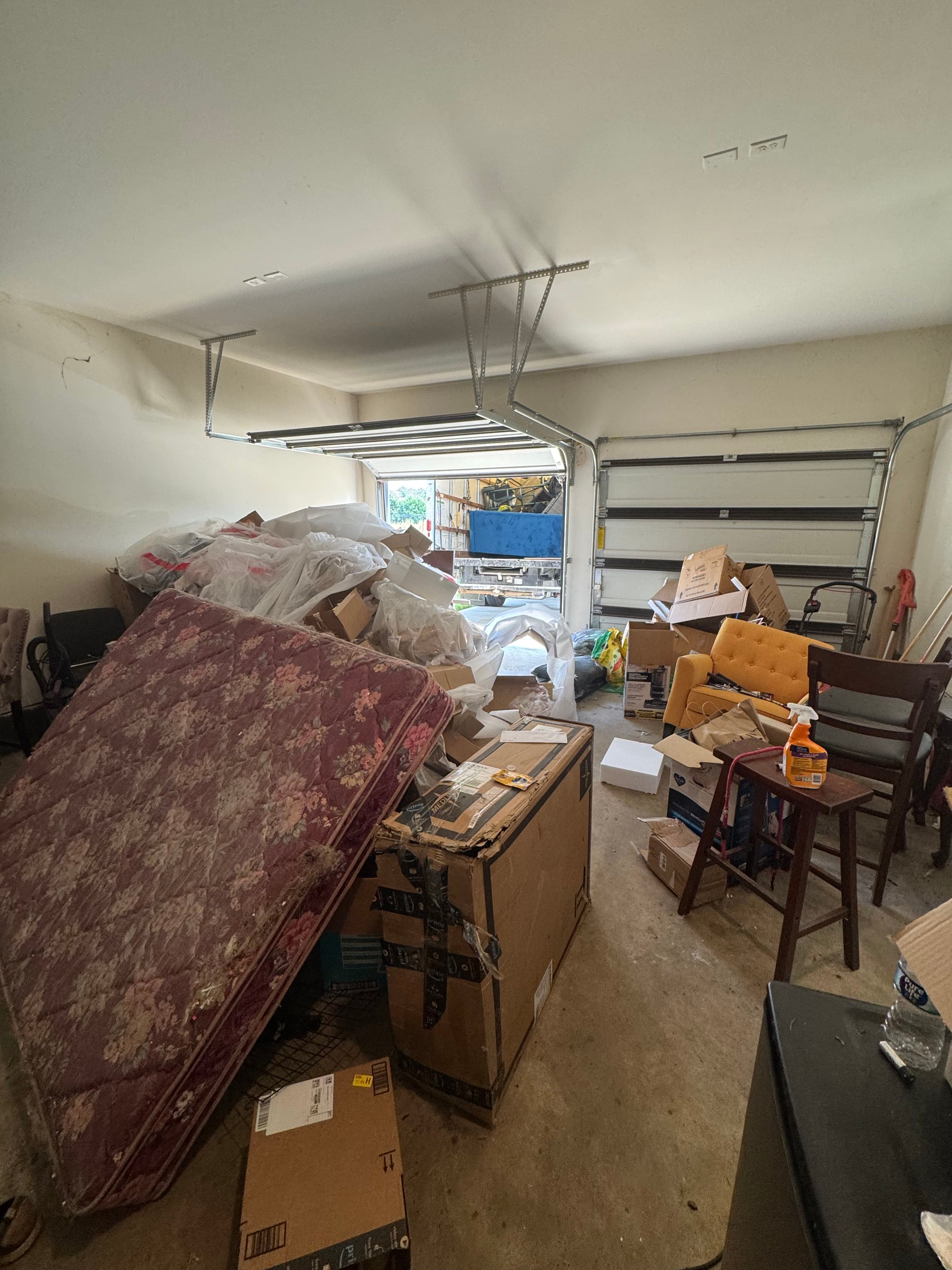 Packed garage before junk removal with a stained mattress, stacked boxes, chairs, and loose items blocking the space.